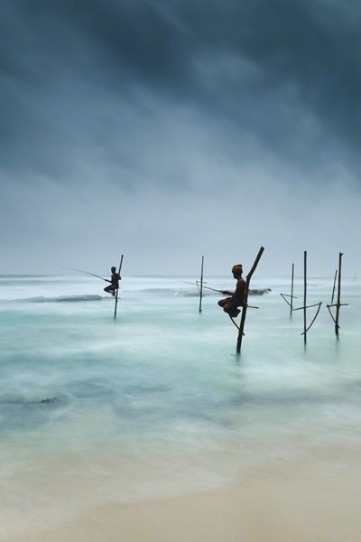 Fishermen catching small reef fish by sitting on small benches attached to poles which are stuck into the water a few meters offshore, known locally as stilt fishing.
